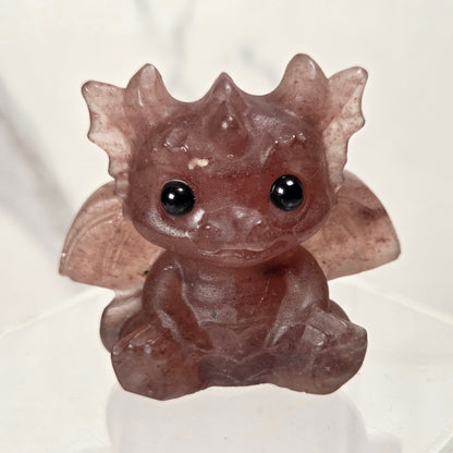 A small translucent brown crystal dragon figurine with large black eyes, sitting on a clear acrylic display stand against a light marble-patterned background.
