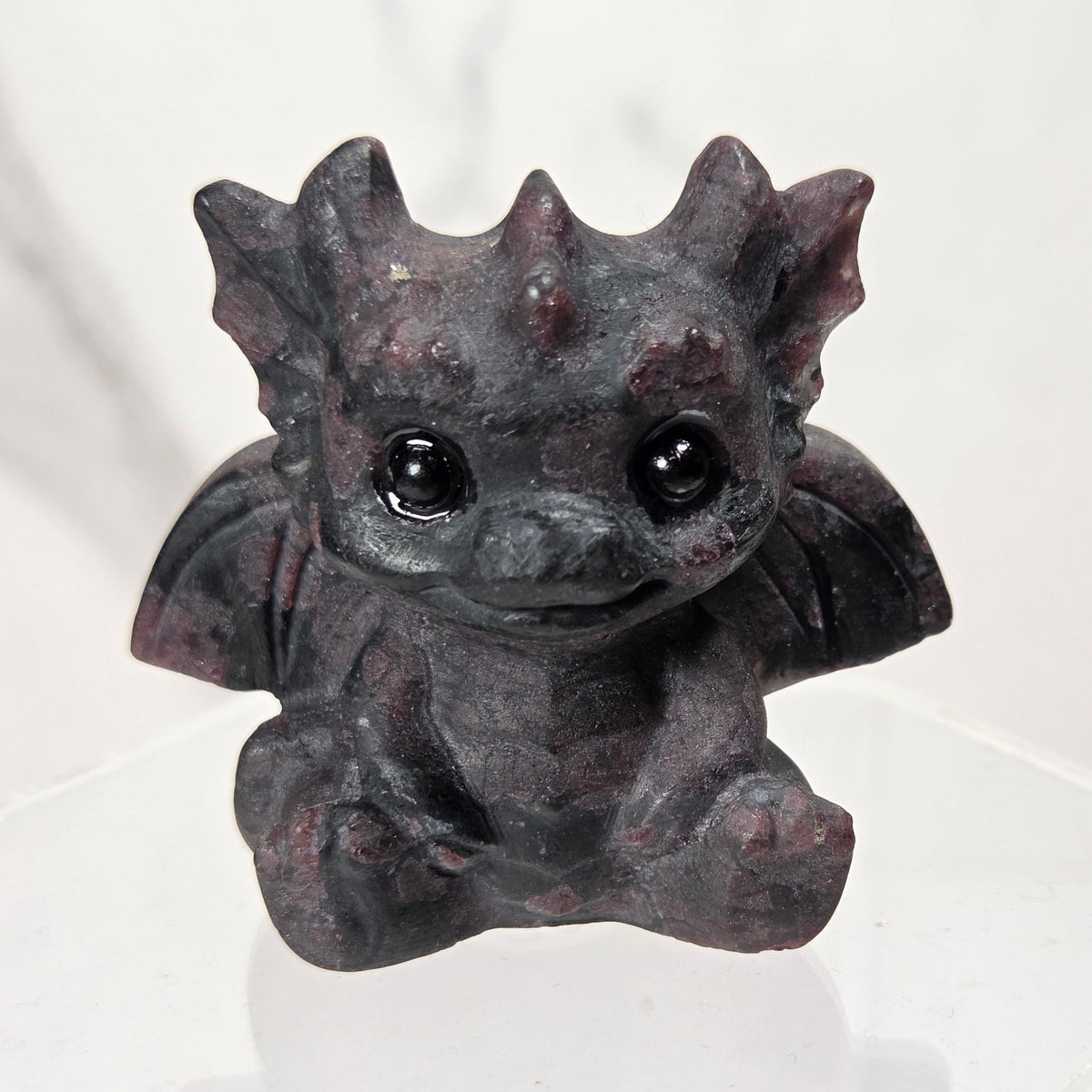 A small black and red crystal dragon figurine with large black eyes, sitting on a clear acrylic display stand against a light marble-patterned background.