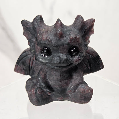 A small black and red crystal dragon figurine with large black eyes, sitting on a clear acrylic display stand against a light marble-patterned background.