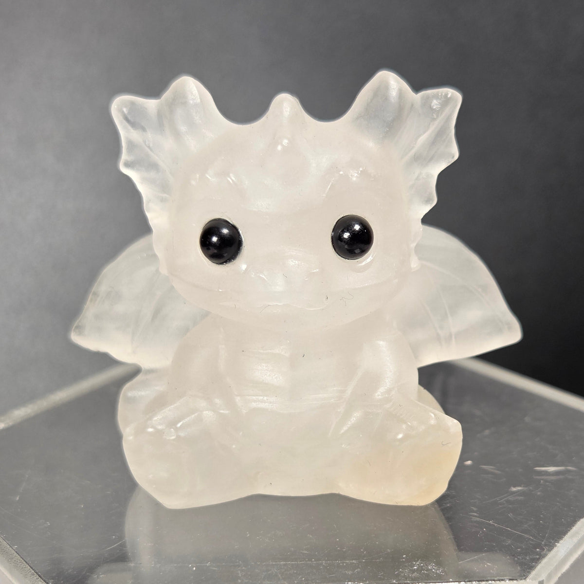 A small translucent crystal dragon figurine with large black eyes, sitting on a clear acrylic display stand against a dark background.