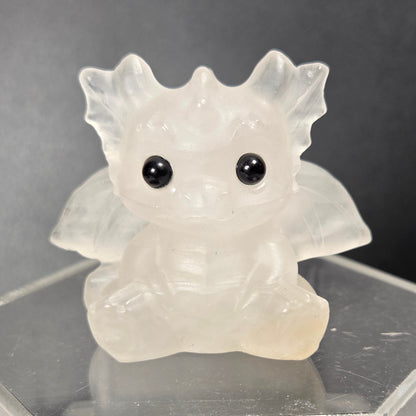 A small translucent crystal dragon figurine with large black eyes, sitting on a clear acrylic display stand against a dark background.