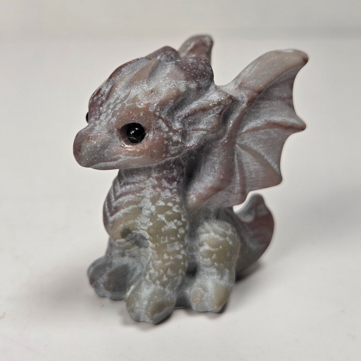 A small gray fantasy dragon figurine with textured scales, wings, and a black eye, standing on a light surface against a soft light gray background.