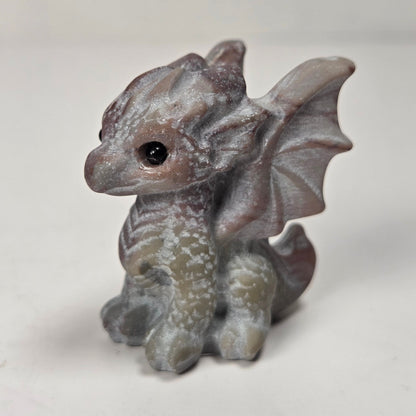 A small gray fantasy dragon figurine with textured scales, wings, and a black eye, standing on a light surface against a soft light gray background.