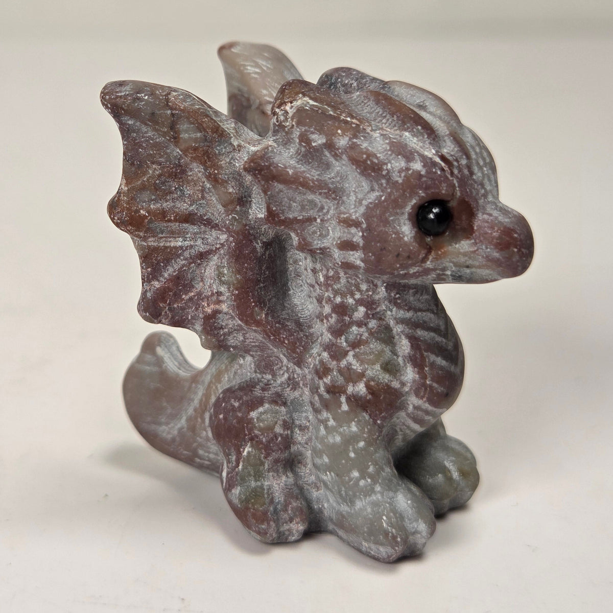 A small gray fantasy dragon figurine with textured scales, wings, and a black eye, standing on a light surface against a soft light gray background.