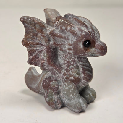 A small gray fantasy dragon figurine with textured scales, wings, and a black eye, standing on a light surface against a soft light gray background.