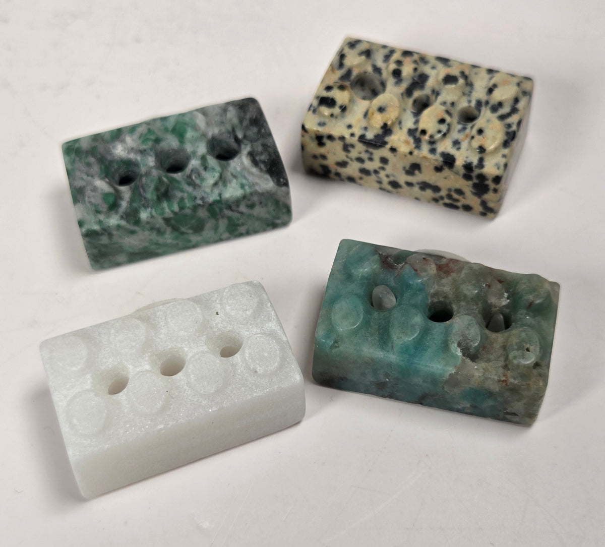 Four rectangular stone beads with holes, displayed on a light off-white background. The beads feature natural stone patterns in green, black-and-beige speckled, solid white, and teal-green.
