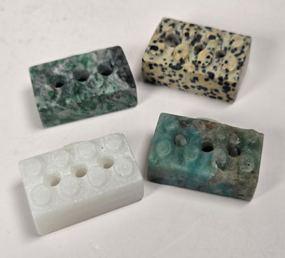 Four rectangular stone beads with holes, displayed on a light off-white background. The beads feature natural stone patterns in green, black-and-beige speckled, solid white, and teal-green.