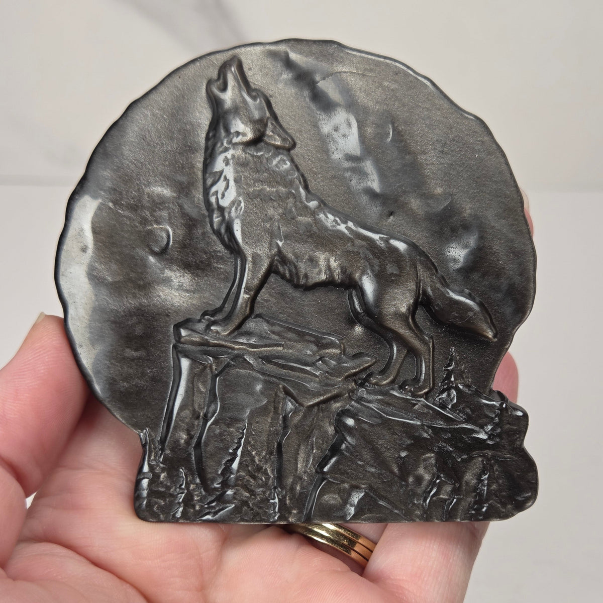 A dark gray cast metal carving featuring a detailed relief of a howling wolf on a rocky cliff, held in a person's hand against a light gray marble background.
