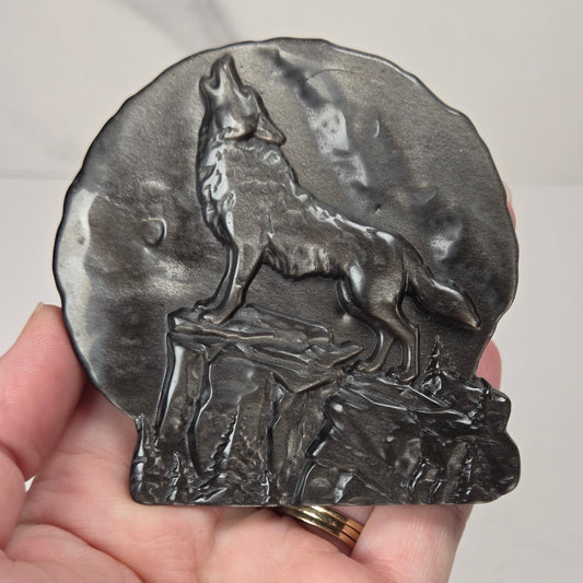 A dark gray cast metal carving featuring a detailed relief of a howling wolf on a rocky cliff, held in a person's hand against a light gray marble background.