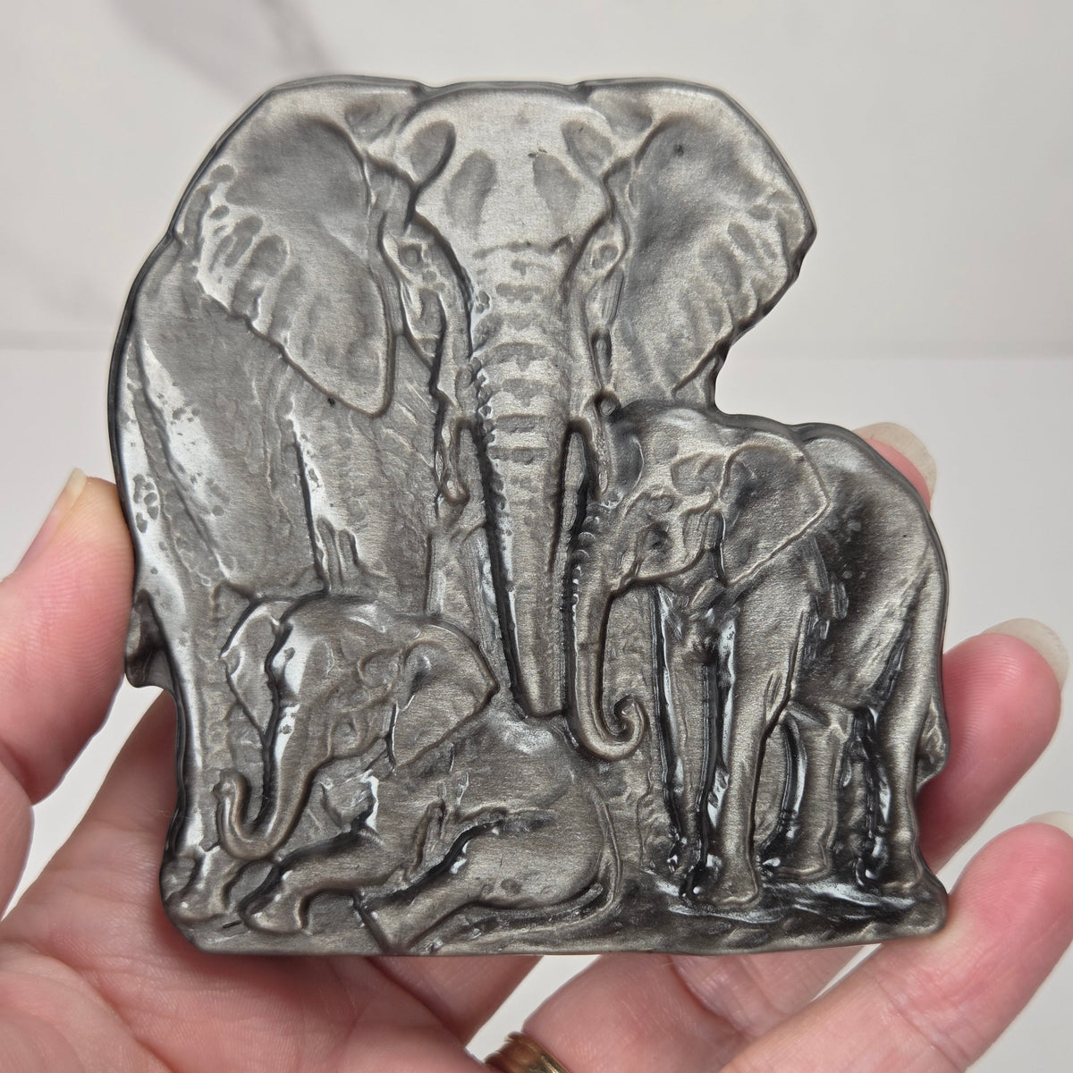 A dark gray cast metal carving featuring a detailed relief of a family of elephants, held in a person's hand against a light gray marble background.