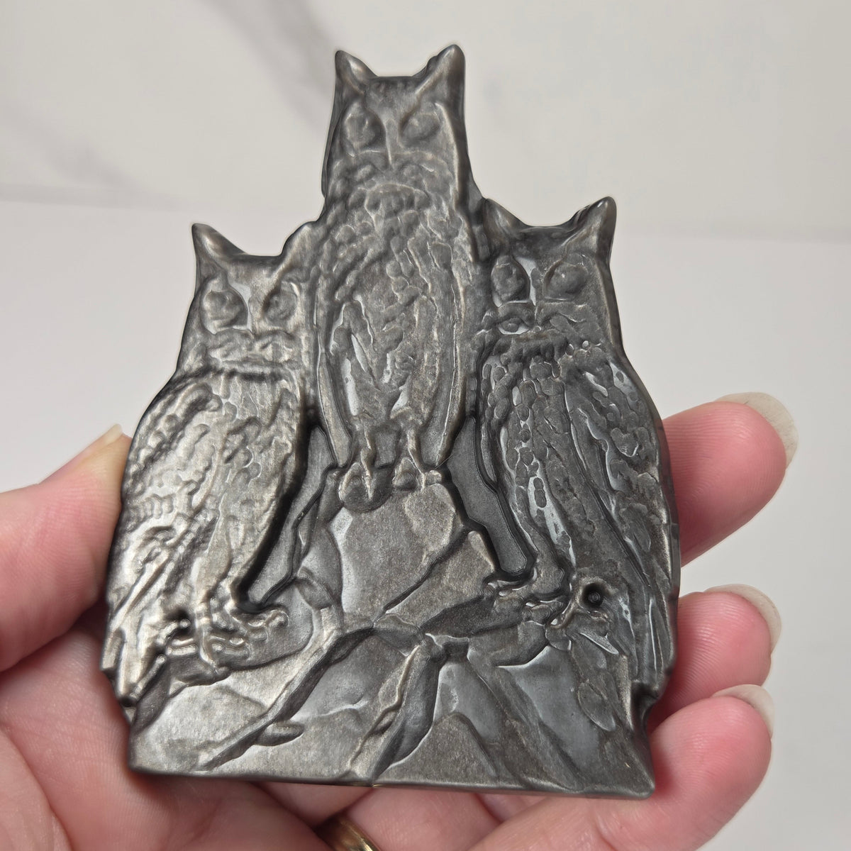 A dark gray cast metal carving featuring a detailed relief of a family of owls, held in a person's hand against a light gray marble background.