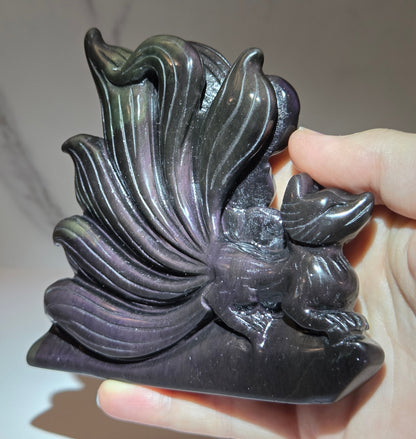 Obsidian stylized fox carving with a light shining highlighting purple coloring being held in a hand against a white marbled background.