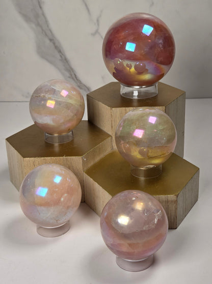 Five iridescent crystal spheres in pink, peach, and reddish tones displayed on clear stands atop gold-colored hexagonal pedestals against a light marble-patterned background.