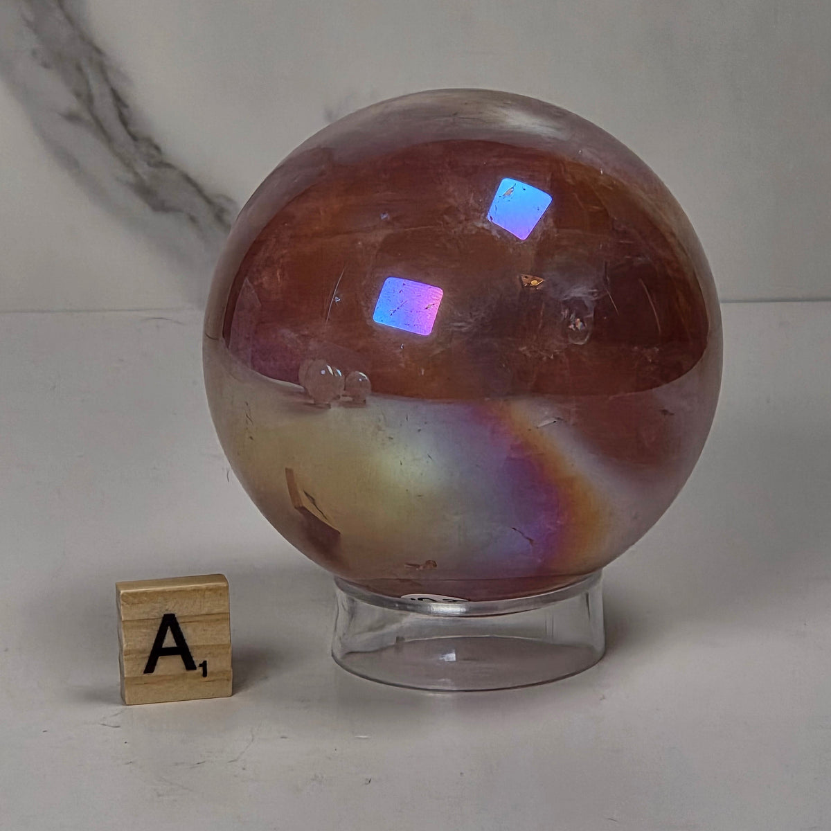  iridescent crystal sphere in pink, peach, and reddish tones displayed on clear stands atop gold-colored hexagonal pedestals against a light marble-patterned background.