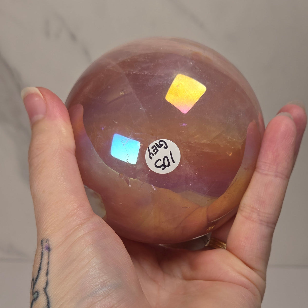 A hand holds a polished round crystal sphere with warm amber-brown tones and iridescent rainbow reflections, against a light marble-look wall background.