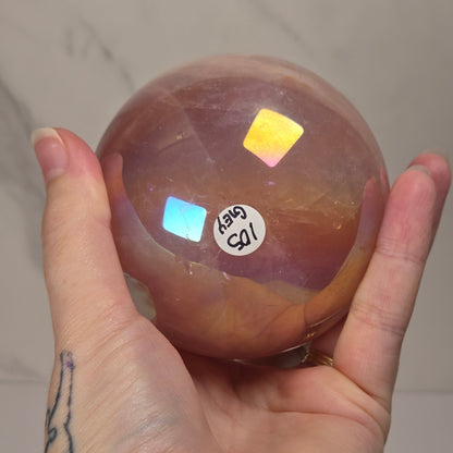 A hand holds a polished round crystal sphere with warm amber-brown tones and iridescent rainbow reflections, against a light marble-look wall background.