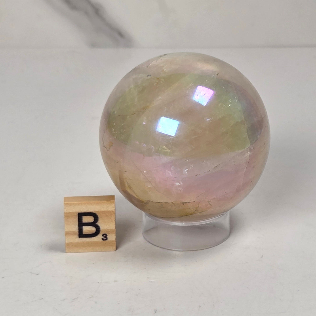 An iridescent crystal sphere with pastel pink, green, and gold tones resting on a clear acrylic stand, next to a small Scrabble tile showing the letter B.