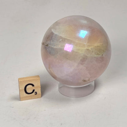 A pink iridescent crystal sphere on a clear display stand, shown next to a small wooden Scrabble tile with the letter C on a white surface.