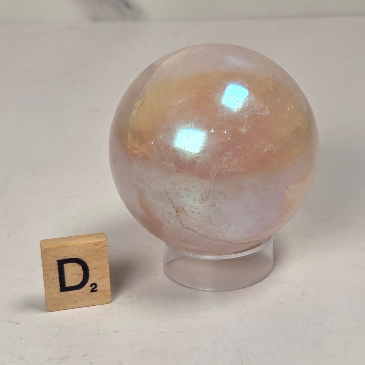 An iridescent peach and cream quartz crystal sphere on a clear acrylic stand, shown next to a wooden Scrabble tile with the letter D2 on a light gray marble-patterned surface.