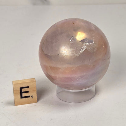 A polished pinkish-tan crystal sphere on a clear display stand, photographed on a light surface with a marble-look background. A small wooden letter tile with the letter E is placed beside it for scale.
