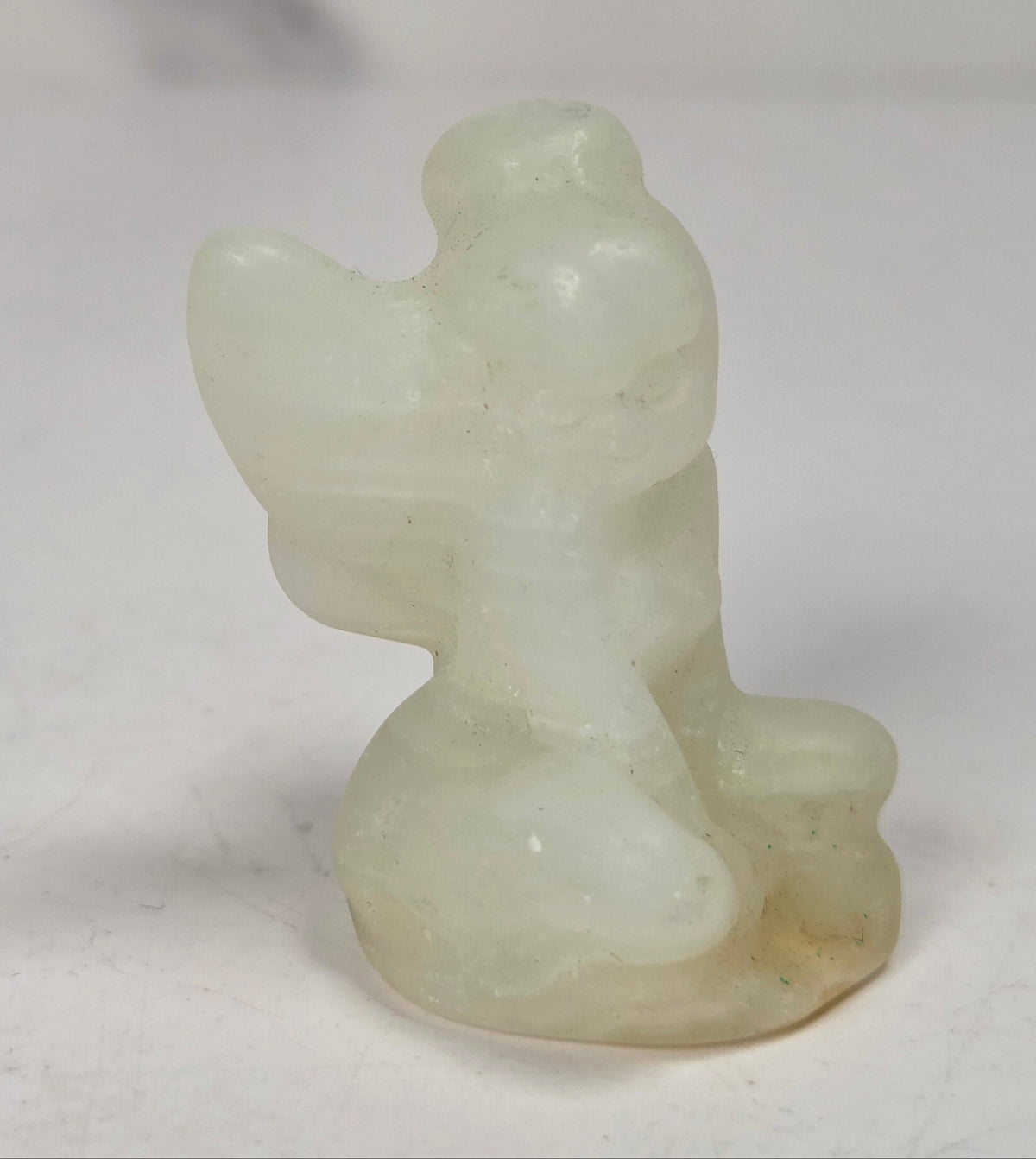 A small carved figurine of a seated angel or cherub, made from pale translucent stone with a soft off-white and light greenish cast, photographed on a plain light background.