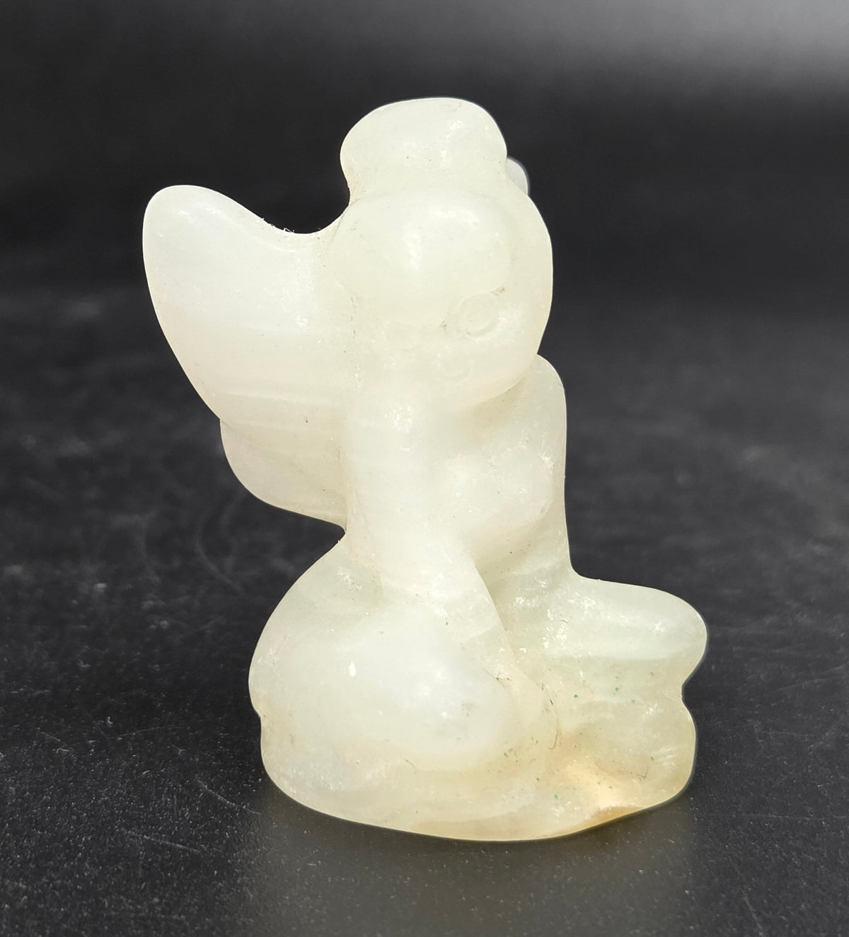 A small carved figurine of a seated angel or cherub, made from a pale translucent stone or resin, photographed on a dark black background.