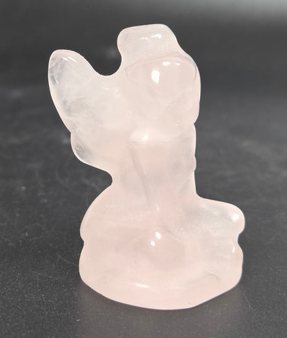 A small translucent pale pink carved figurine or crystal ornament standing on a dark gray surface against a dark gray background.