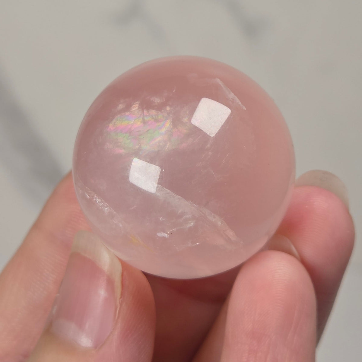 A hand holding a smooth, polished pink stone sphere with a soft iridescent sheen, set against a light gray marble-patterned background.