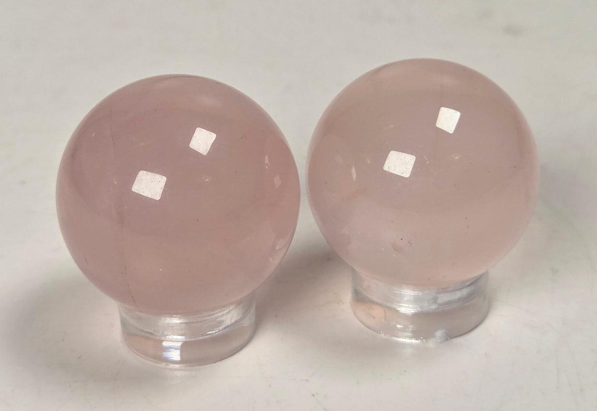 Two smooth, polished pale pink stone spheres on clear acrylic stands, photographed on a light neutral surface.