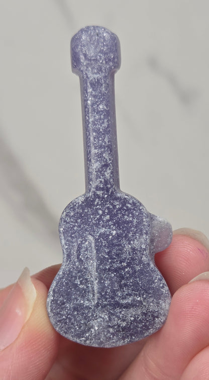 A miniature purple guitar-shaped stone carving held between fingers against a light gray marble background.
