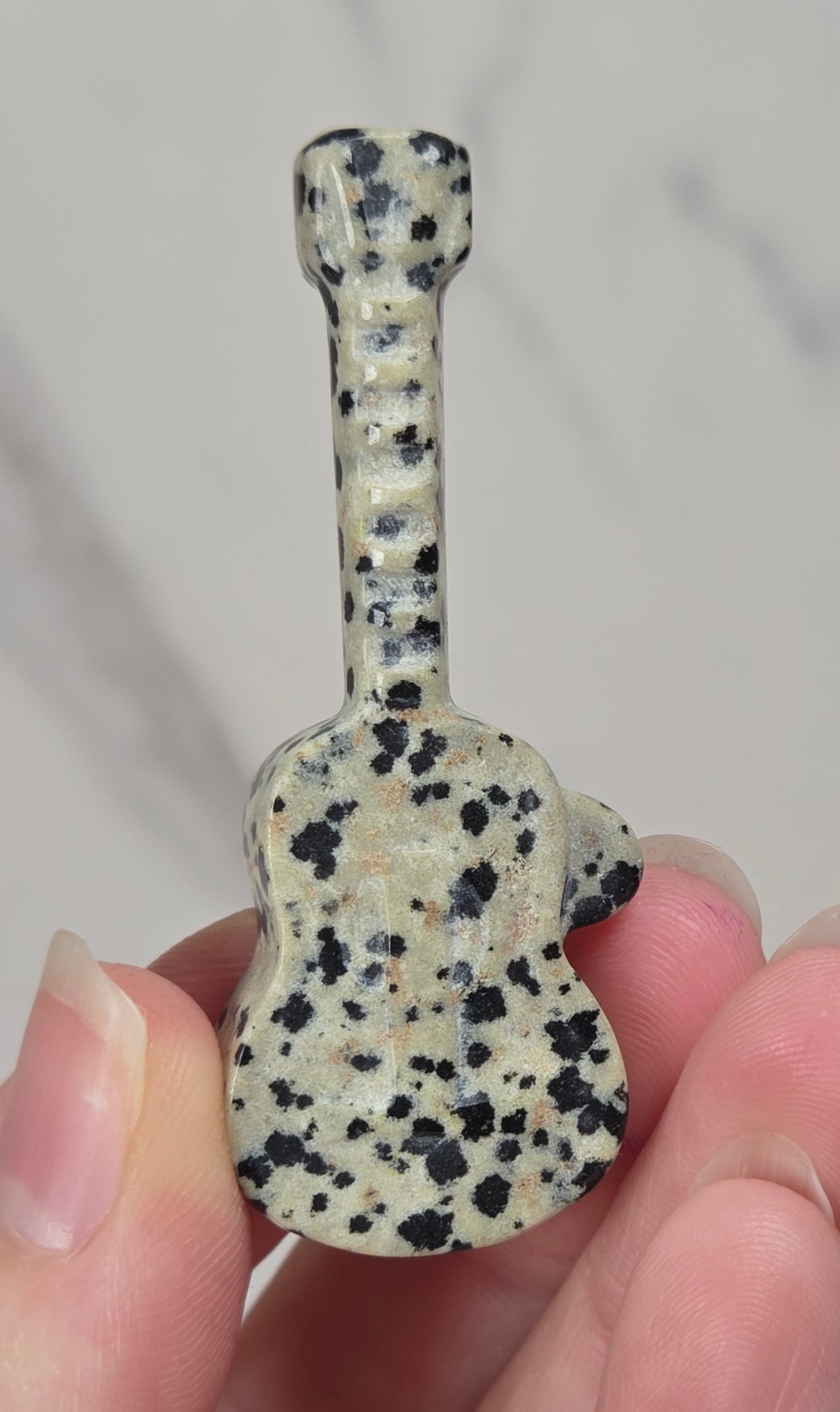 A small guitar-shaped pendant or charm made of speckled stone, held between fingers against a light gray marble-like background.