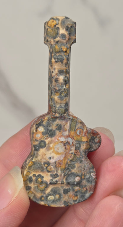A miniature guitar-shaped charm or pendant with a mottled, textured pattern of brown, gold, and black spots, held in a person's hand against a light gray marble background.