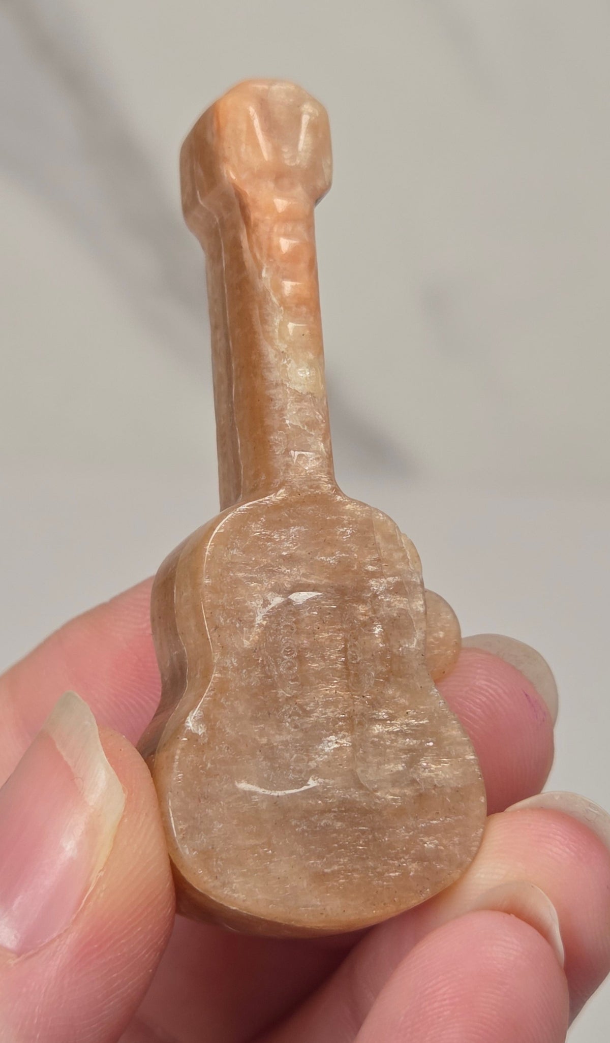 A hand holding a small carved guitar-shaped object made of pale peachy-brown stone or resin, photographed against a soft light gray background.