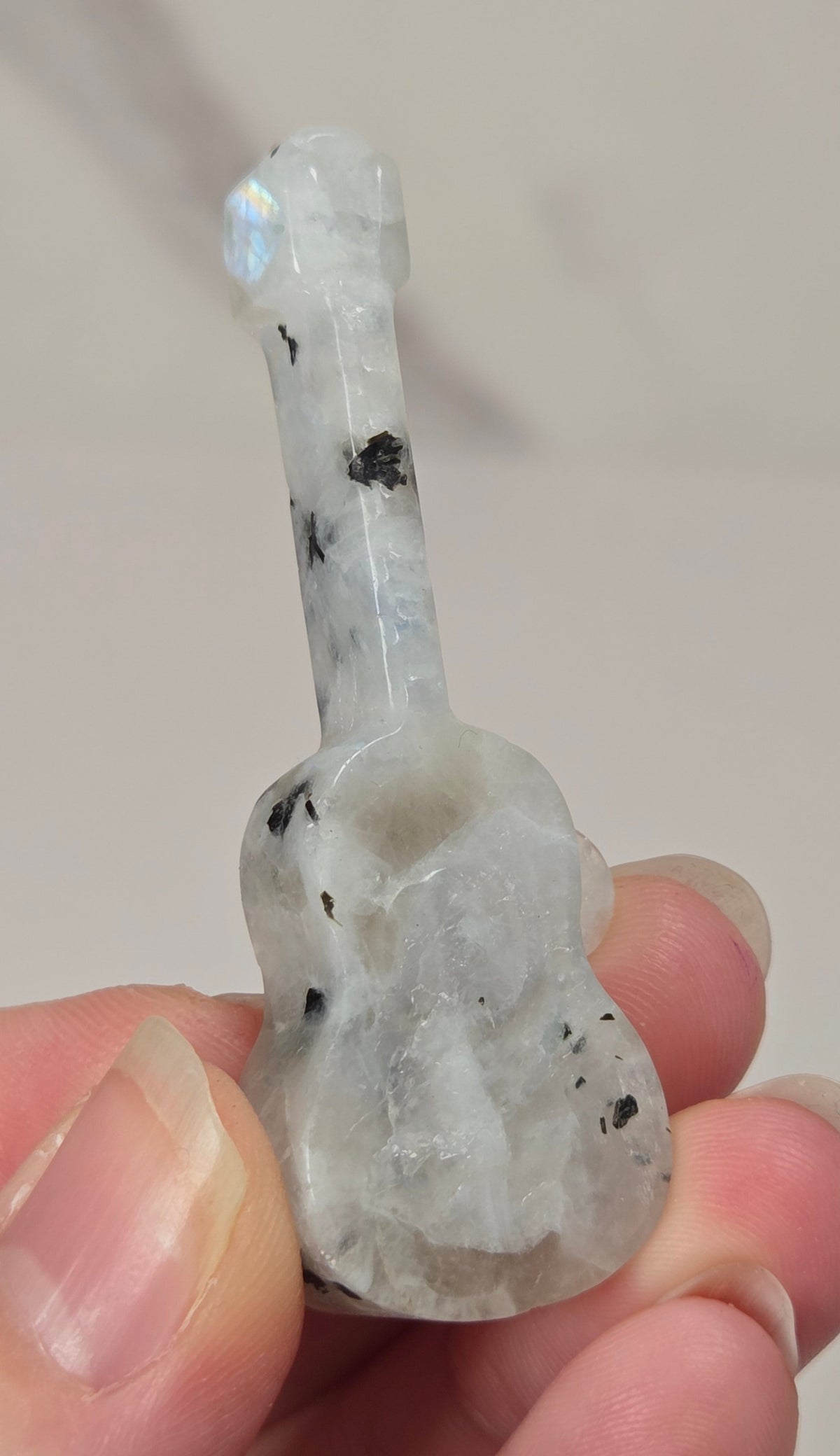 A hand holding a small carved guitar-shaped object made of milky translucent stone with black inclusions and a subtle iridescent blue flash near the neck.