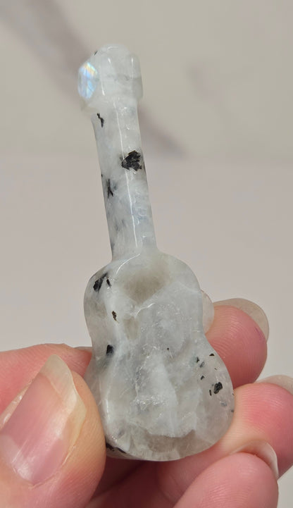 A hand holding a small carved guitar-shaped object made of milky translucent stone with black inclusions and a subtle iridescent blue flash near the neck.