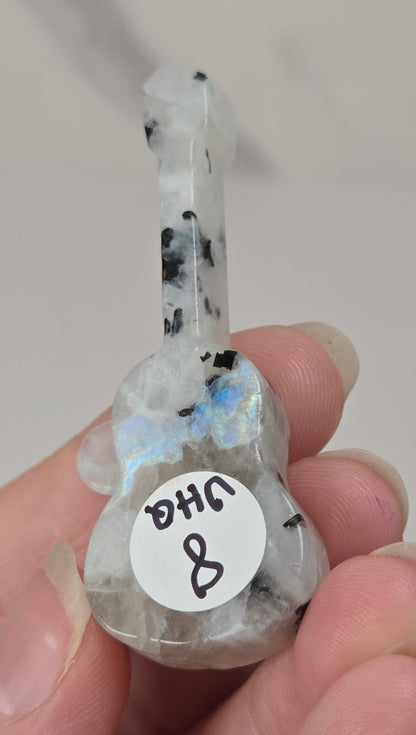 A hand holding a small carved gemstone guitar-shaped pendant or charm with black speckles and a blue iridescent sheen, featuring a white circular sticker with handwritten text.