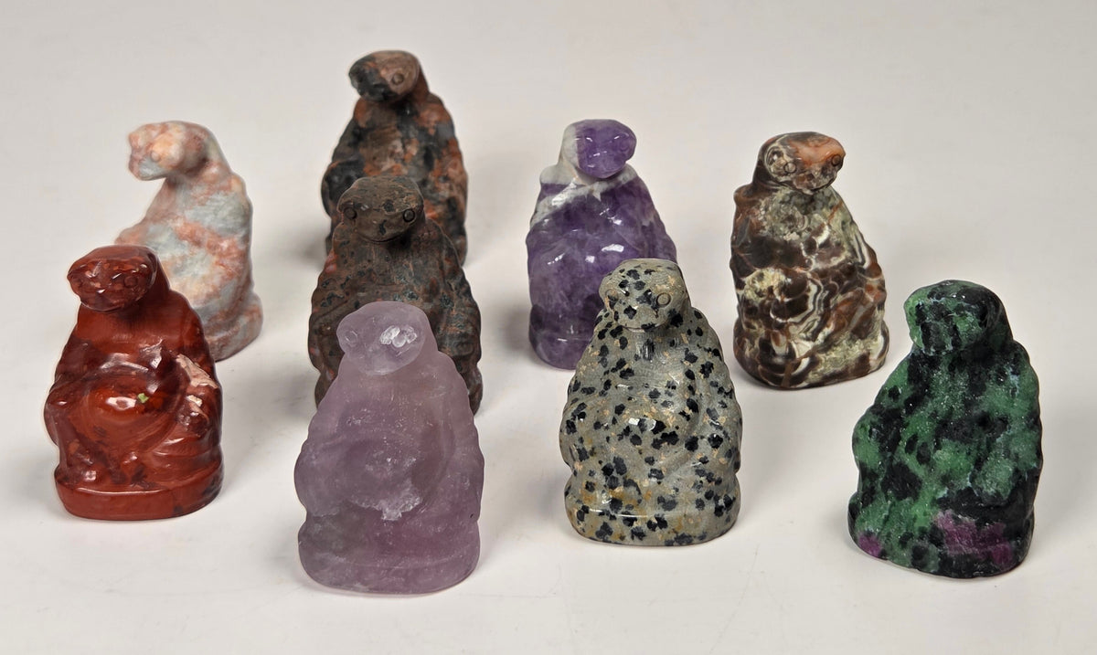 A set of eight small hand-carved crystal figurines shaped like seated figures, displayed on a plain light background. The figures are made from various colored stones, including red, purple, green, gray, and brown.