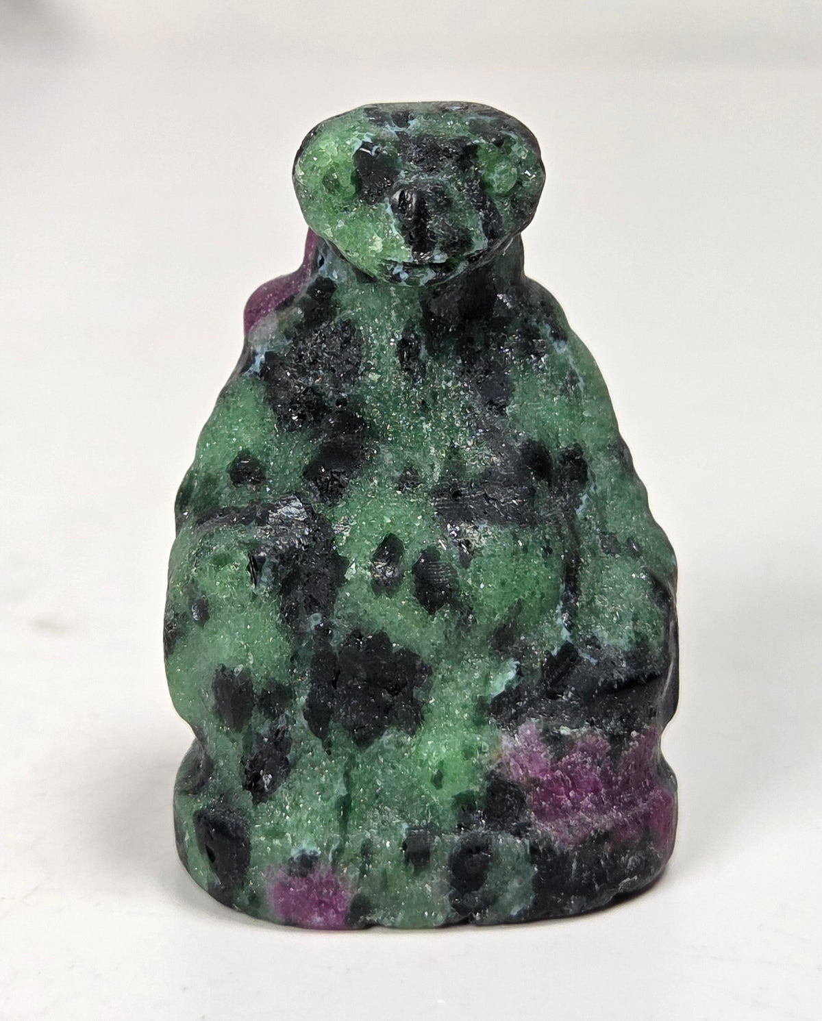 A small carved figurine of a seated figure, likely a monkey or primate, made from green and black mottled stone with hints of purple, photographed on a plain light background.