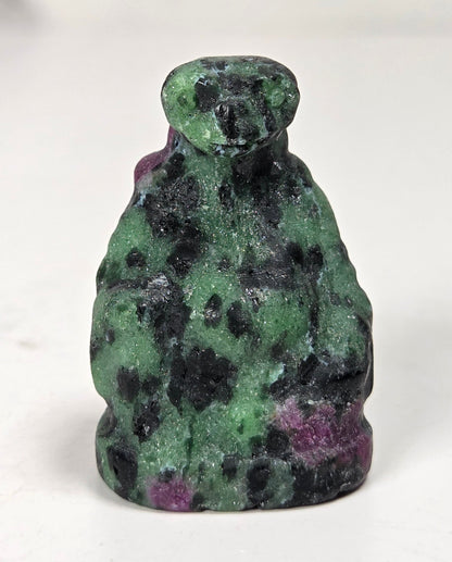 A small carved figurine of a seated figure, likely a monkey or primate, made from green and black mottled stone with hints of purple, photographed on a plain light background.