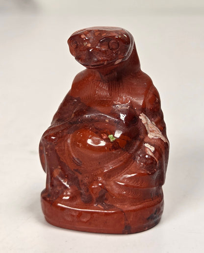A small carved figurine of a seated monkey, made from reddish-brown polished stone or resin, set against a plain off-white background.