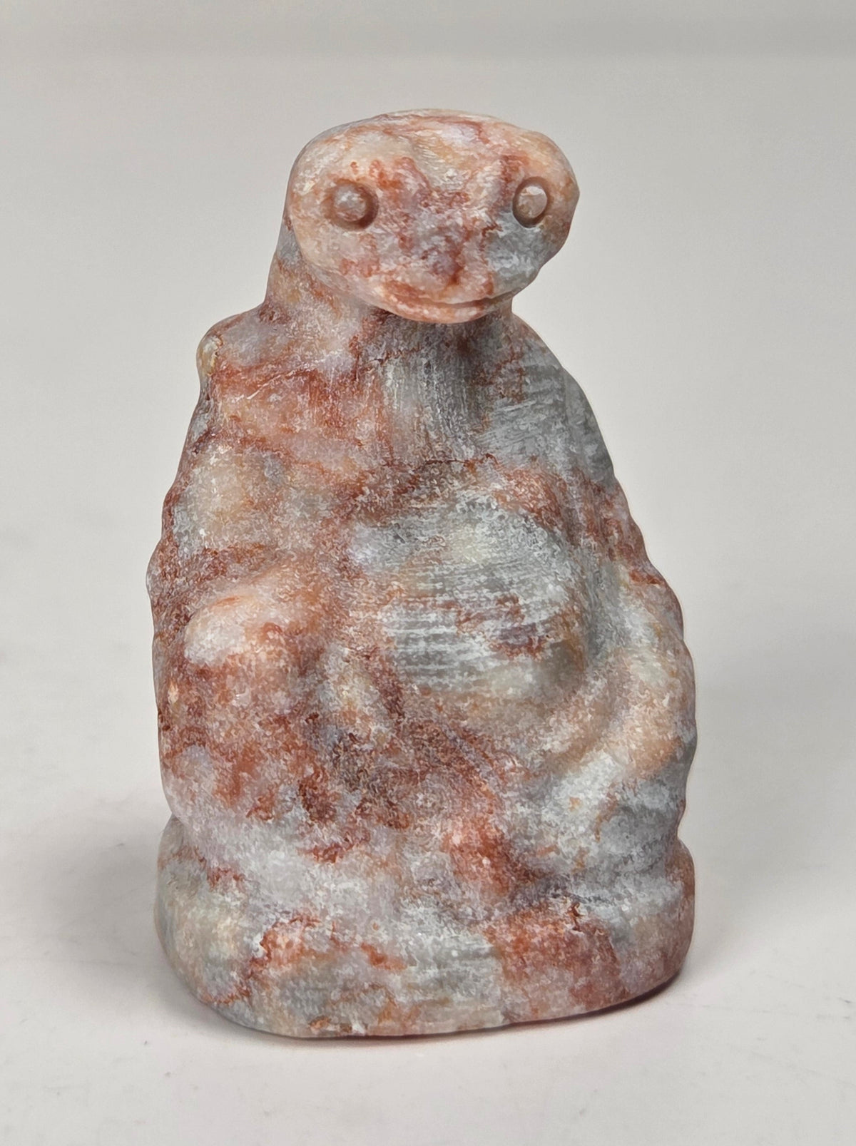 A small carved figurine of a seated figure, likely made of stone or resin, featuring a mottled coloration of reddish-brown, gray, and off-white. The figurine is photographed on a neutral light gray surface.
