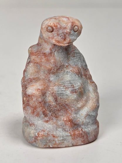 A small carved figurine of a seated figure, likely made of stone or resin, featuring a mottled coloration of reddish-brown, gray, and off-white. The figurine is photographed on a neutral light gray surface.