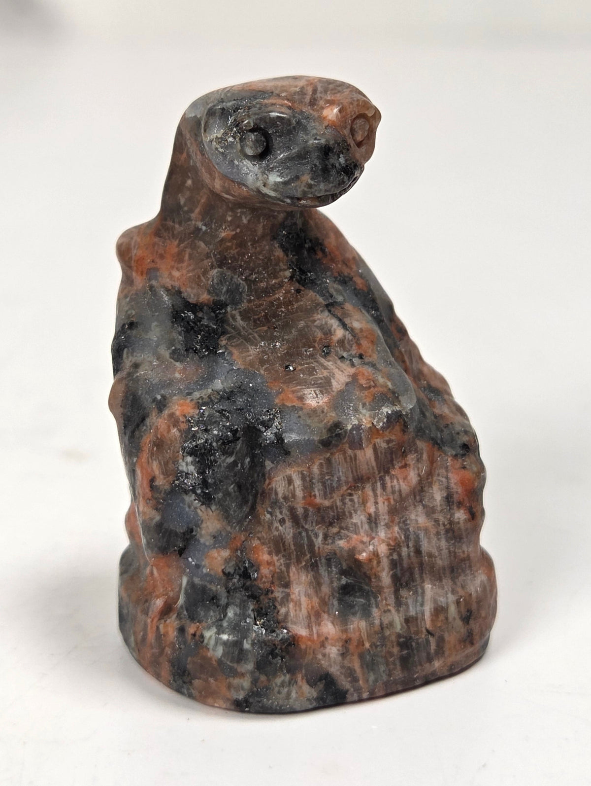 A small carved figurine resembling a seated figure or animal head, made from mottled stone with dark charcoal and rust-brown tones, photographed on a plain light background.