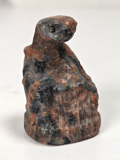 A small carved figurine resembling a seated figure or animal head, made from mottled stone with dark charcoal and rust-brown tones, photographed on a plain light background.