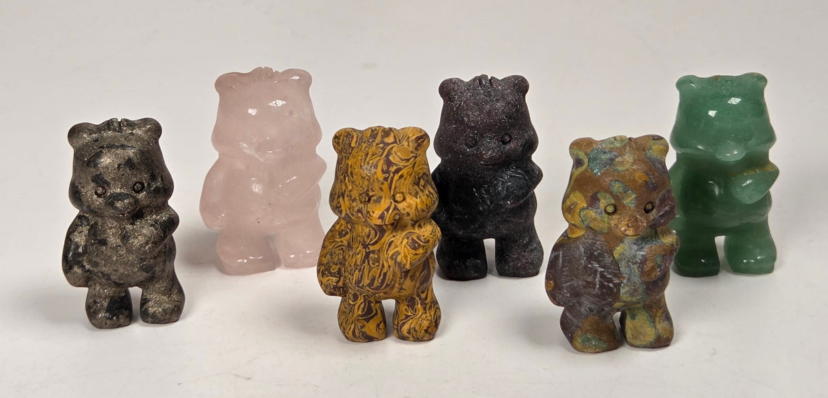 Five small carved bear figurines in different stone colors and patterns, including gray, pale pink, yellow-brown, dark charcoal, and green, arranged on a white marble-like surface.