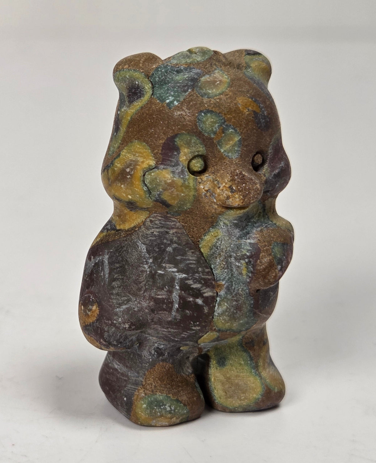A small figurine of a bear wearing a scarf, featuring a textured patina with brown, green, and gold tones, set against a plain light gray background.