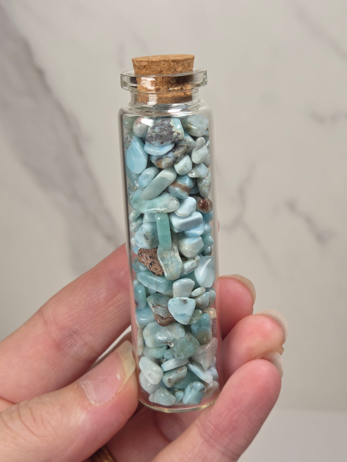 A hand holding a small clear glass vial with a cork stopper filled with polished light blue stone chips, set against a light marble background.