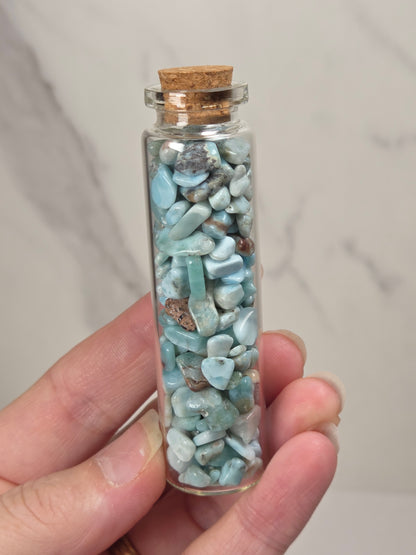 A hand holding a small clear glass vial with a cork stopper filled with polished light blue stone chips, set against a light marble background.