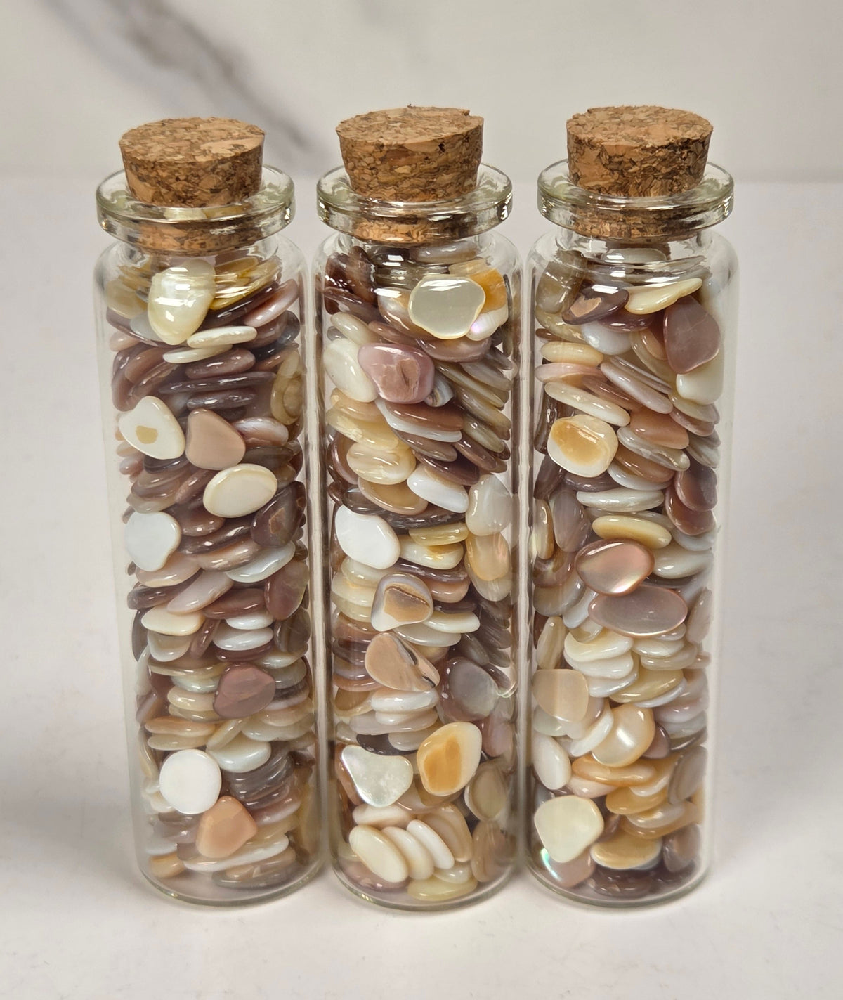 Three clear glass vials with cork stoppers filled with small flat decorative pieces in brown, cream, and beige tones, arranged on a light surface against a soft marble-like background.