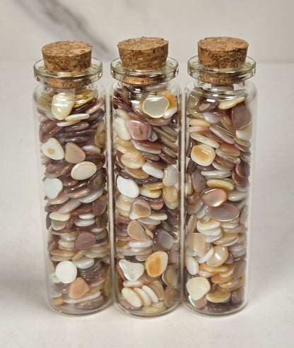 Three clear glass vials with cork stoppers filled with small flat decorative pieces in brown, cream, and beige tones, arranged on a light surface against a soft marble-like background.